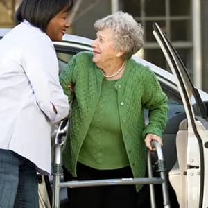 A caregiver steadies a smiling senior using a walker as she steps out of a vehicle.