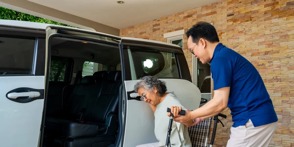A caregiver in a blue shirt gently helps a smiling senior with a cane step into a clean, accessible van.